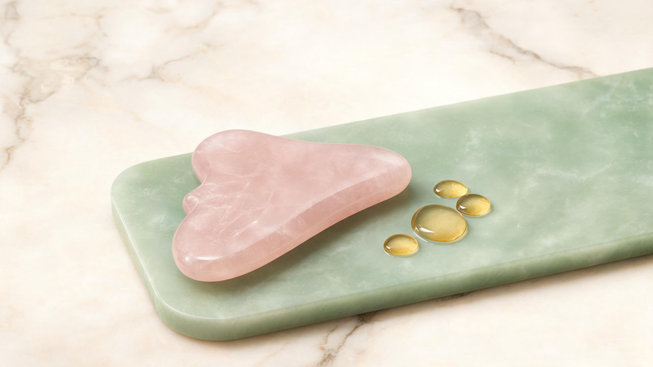 A rose quartz gua sha tool resting on a jade-green surface with drops of golden facial oil